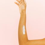 A close-up of a skincare routine with lotion on an arm, set against a pink backdrop.