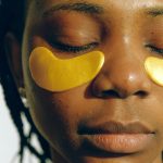 Crop gentle African American female with closed eyes and clean face enjoying cosmetic procedure for smoothing and moisturizing skin under eyes and removing dark circles