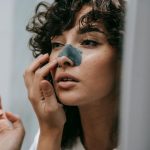 Mirror reflection of young female with curly hair putting nose strip mask on face in bathroom